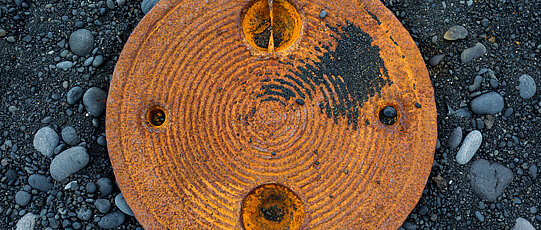 Round, rusty metal piece with decorative grooves on black sand and pebbles.