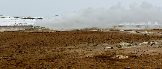 Smoking geyser on brown ground with snowy hills in the background.