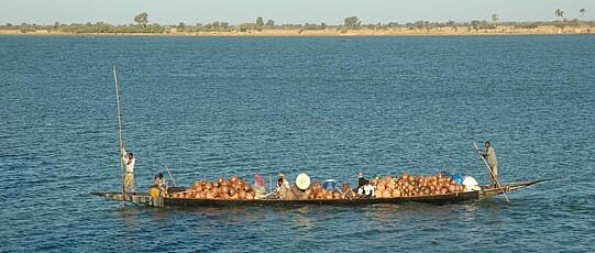 Vollbeladenes, langes Boot, das von zwei Leuten gelenkt wird; im Hintergrund Ufer.