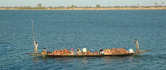 Fully loaded, long boat, steered by two people; in the background shore.