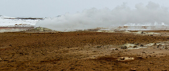 Rauchender Geysir auf braunem Boden mit schneebedeckten Hügeln im Hintergrund.